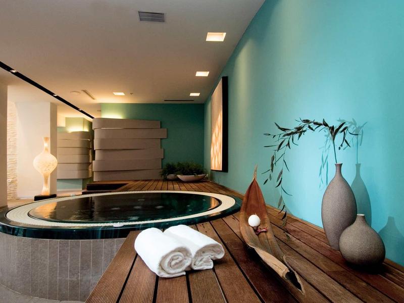 Moderní wellness se okrouhlou vanou, dřevěnými lavicemi a dekorativními vázami na modré zdi.
