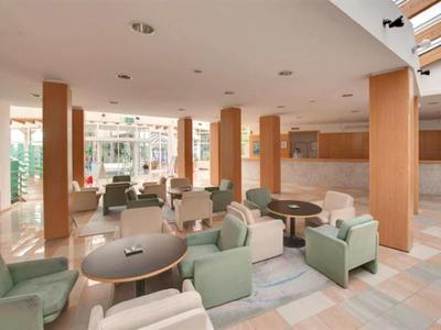 Gemütlicher Hotel-Loungebereich mit Tischen, Sesseln und großen Fenstern für viel Tageslicht.