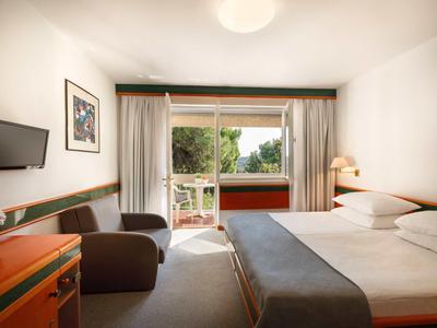 Helles Hotelzimmer mit Doppelbett, Sesseln, Fernseher und Balkon mit Blick auf Bäume.