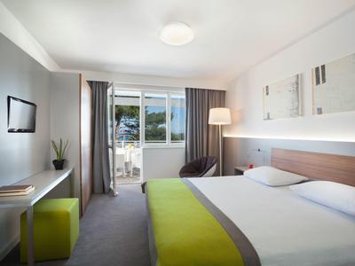 Helles Hotelzimmer mit Doppelbett, modernem Schreibtisch und Balkon mit Blick auf Wasser.