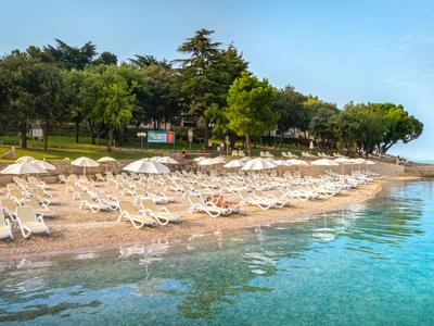 Strand mit Liegestühlen und Sonnenschirmen neben klarem, ruhigem Wasser und bewachsenen Hügeln.