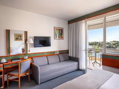 Helles Hotelzimmer mit grauem Sofa, Schreibtisch, TV und Balkon mit Blick ins Grüne.