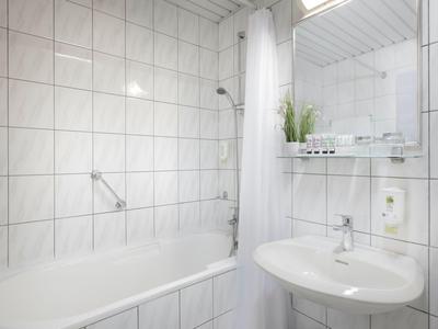 Weiß gefliestes Badezimmer mit Badewanne, Duschvorhang, Waschbecken und Regal mit Pflanze.