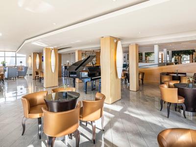 Heller Hotel-Loungebereich mit runden Tischen, braunen Sesseln und Klavier im Hintergrund.