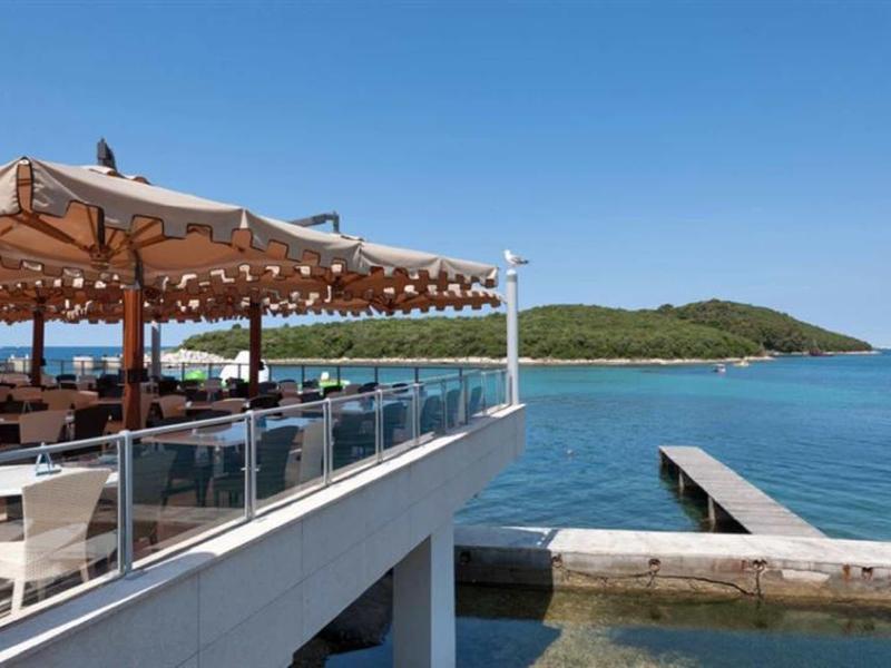 Terasa restaurace u moře s deštníky s výhledem na mol a kopce.