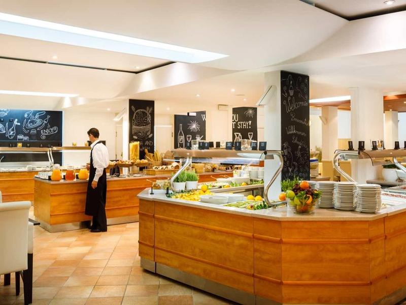 Moderní hotelová restaurace s dřevěnými pulty, bílými židlemi a bufetovou částí.