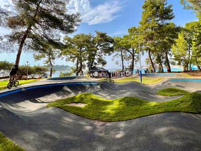 Park s členitým travnatým terénem a cyklisty pod stromy za slunečného dne