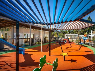 Un parque infantil soleado con tobogán y columpios bajo una protección solar azul y blanca.