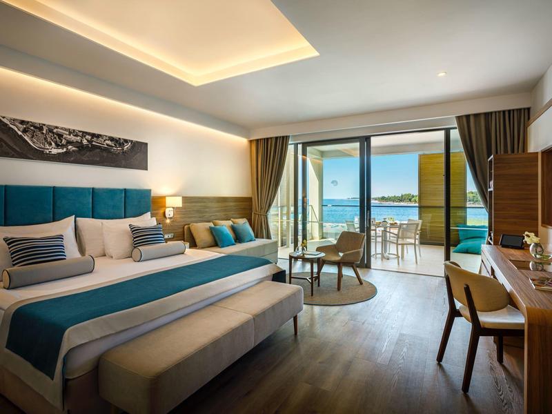 Habitación de hotel moderna con cama grande, escritorio, balcón y vista al mar a la luz del día.