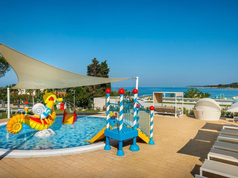 Área de piscina infantil con juegos de agua y tumbonas bajo un cielo azul despejado.
