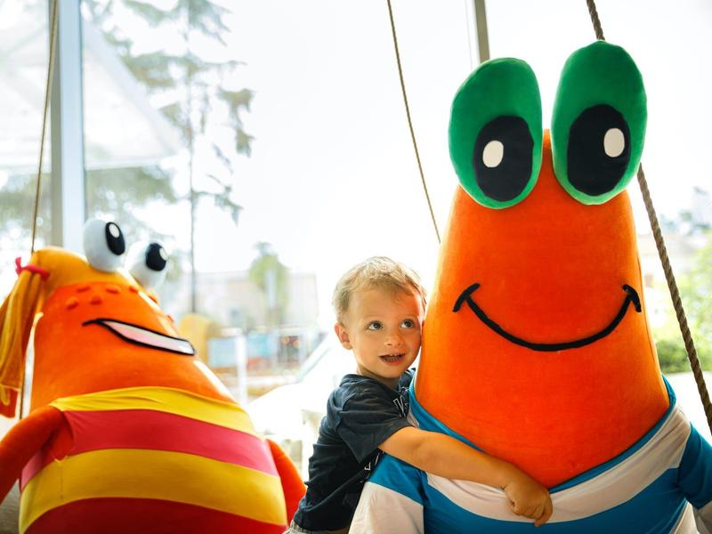 Un niño abraza dos peluches grandes y coloridos con caras amigables en un interior.