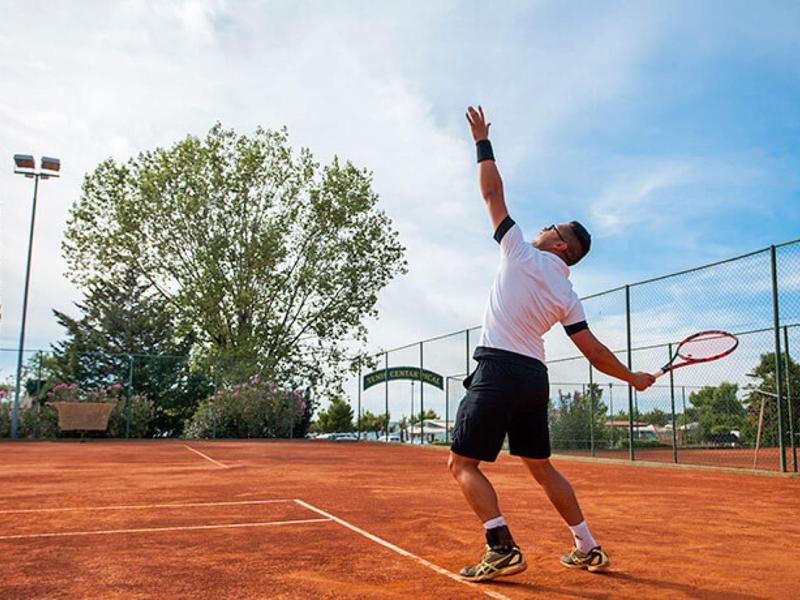 Una persona juega al tenis en una pista de arcilla roja bajo un cielo nublado, rodeada de árboles y una cerca.