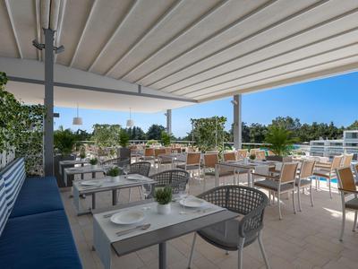Terrasse avec tables, chaises et bancs sous un grand auvent, entourée de verdure.