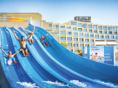 Kilka osób zjeżdża na niebieskich zjeżdżalniach wodnych przed hotelem.