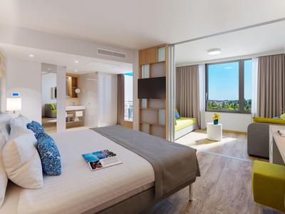 Chambre d'hôtel lumineuse avec grand lit, télévision, et vue sur la ville depuis un coin salon.