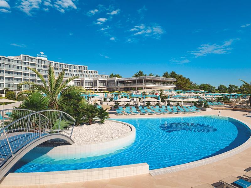 Complexe hôtelier avec grande piscine, chaises longues, palmiers et ciel bleu.