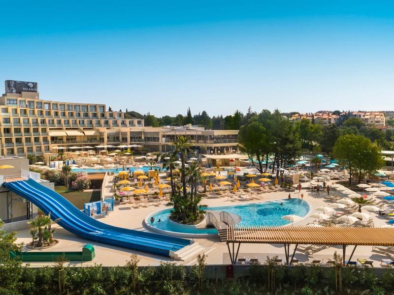 Complexe hôtelier avec piscine, toboggans bleus et terrasses avec chaises longues sous le soleil.