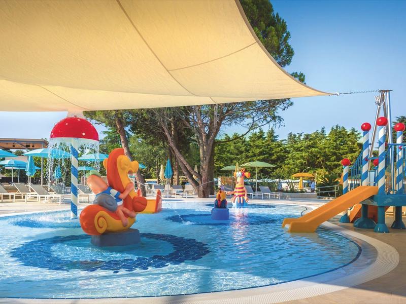 Petite piscine avec jeux d'eau colorés pour enfants, toboggan et parasol dans un parc verdoyant.