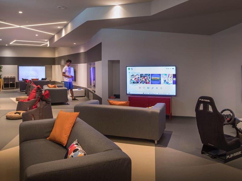 Salon moderne d'hôtel avec canapés, fauteuils de jeu et écrans TV.