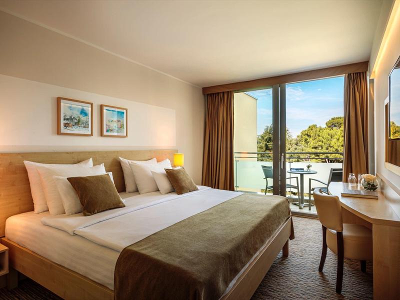 Chambre d'hôtel lumineuse avec lit double, bureau, balcon et vue sur les arbres.