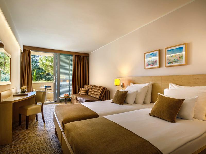 Chambre d'hôtel lumineuse avec deux grands lits, coin salon et balcon avec vue sur le jardin.