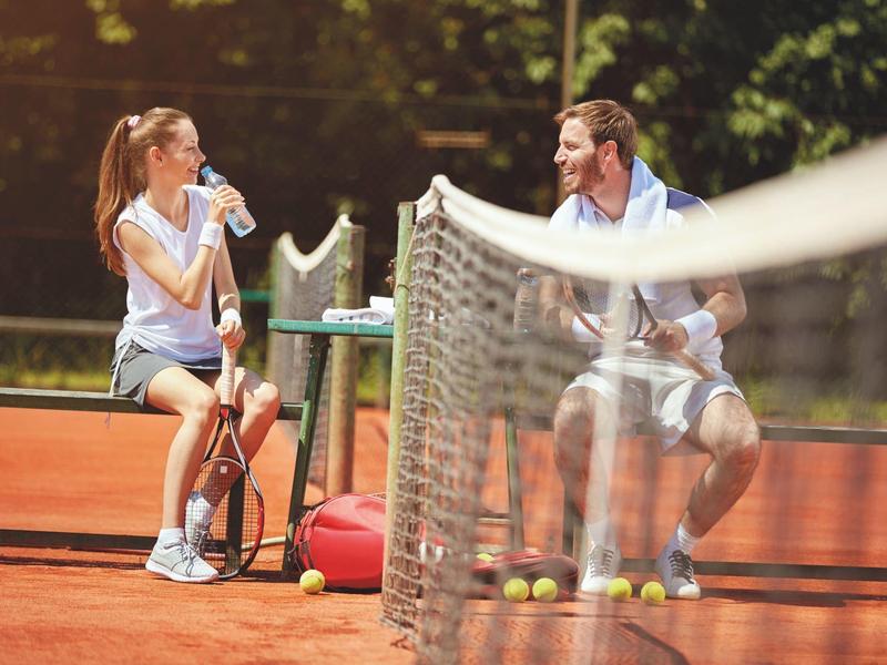Deux personnes reposent sur un banc de tennis, buvant de l'eau, avec une raquette et un sac à côté.