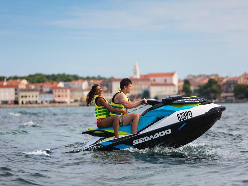 Deux personnes sur un jet ski bleu sur une mer calme avec un village en arrière-plan.