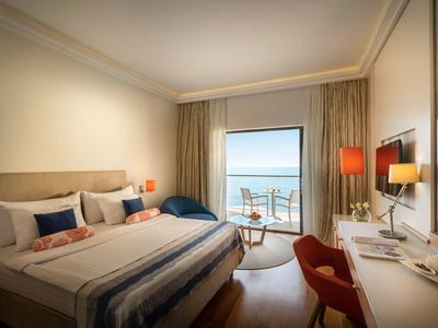 Helles Schlafzimmer mit großem Bett, Balkon mit Meerblick, Schreibtisch, rotem Stuhl und warmem Licht.