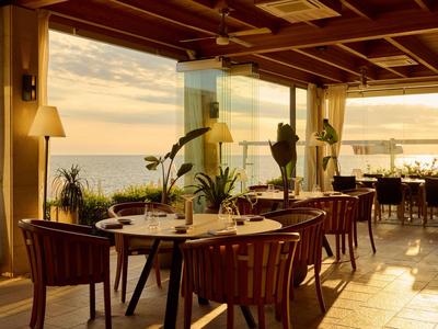 Restaurant mit Holztischen und -stühlen, großen Fenstern, Pflanzen und Blick auf das Meer bei Sonnenuntergang.