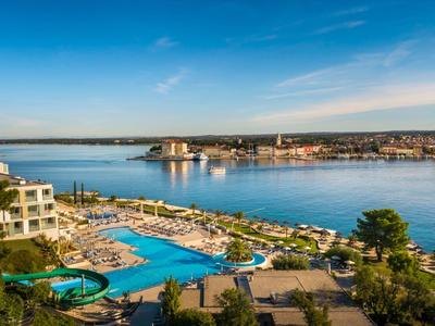 Blick auf ein großes Hotel mit Pool, Sonnenschirmen und Meer im Hintergrund bei klarem Himmel.