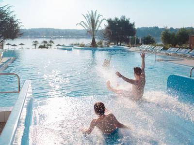 Zwei Personen rutschen im Pool mit Planschwasser und sonnigem Himmel im Hintergrund.