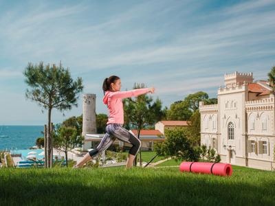 Frau macht Yoga-Übung auf einer Wiese mit Meerblick vor einem Hotelgebäude.