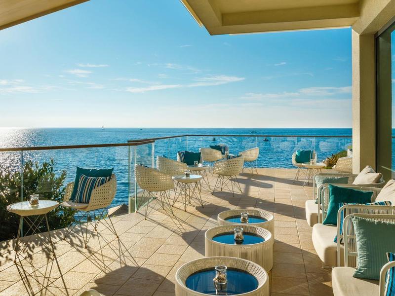 Terrassenbereich mit Sitzmöbeln, Glastüren und Meerblick unter blauem Himmel.