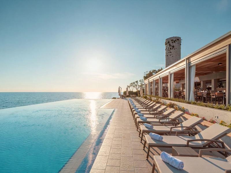 Infinity-Pool mit Sonnenuntergang, links das Meer, rechts Liegestühle und ein langes Gebäude.