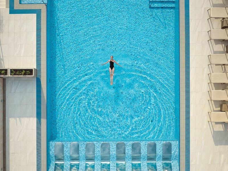 Rechteckiger Pool mit blauen Liegestühlen und einer Person im Becken, umgeben von weißen Liegen.