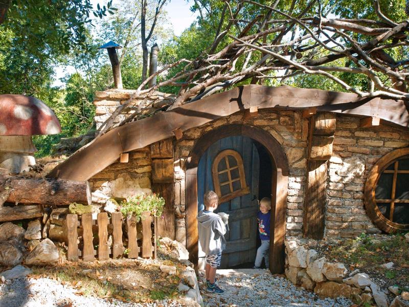 Kleines, rustikales Steinhaus mit rundem Fenster und geschwungener Tür, umgeben von Bäumen.