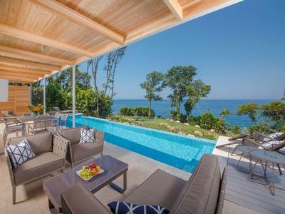 Moderner überdachter Balkon mit Sitzbereich, Pool und Meerblick, umgeben von Bäumen und blauem Himmel.