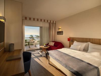 Modern hotel room with double bed, sofa, desk, and view of balcony and sea.
