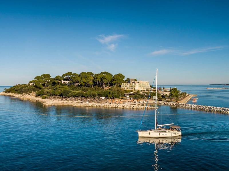 Sailboat near a wooded island with a large building on the shore under clear sky.