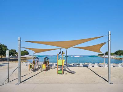 Toldos de sombra sobre un área de juegos de arena en la playa con el mar y el cielo despejado al fondo.
