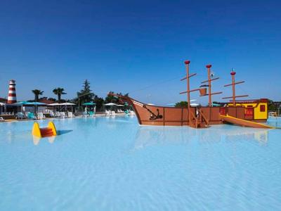 Gran piscina infantil con tobogán en forma de barco pirata y agua azul clara bajo cielo despejado.