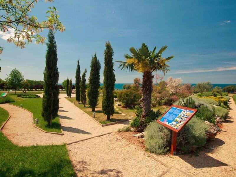 Jardín con caminos de grava, cipreses y palmeras junto al mar bajo un cielo azul claro