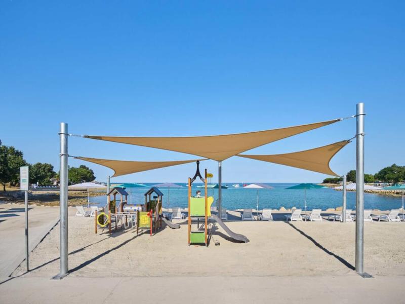 Toldos de sombra sobre un área de juegos de arena en la playa con el mar y el cielo despejado al fondo.