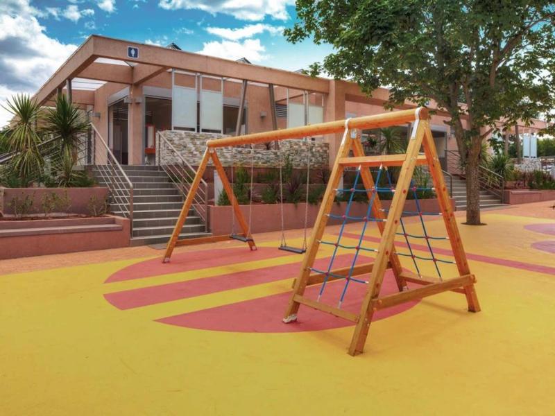 Parque infantil moderno con columpio y edificio de hotel climatizado al fondo bajo el sol.