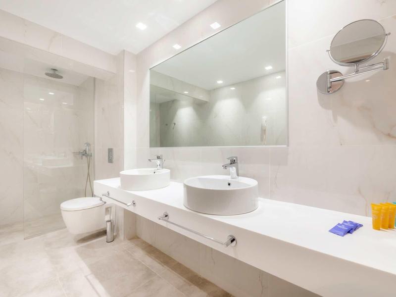 Baño de hotel luminoso con mobiliario blanco, doble lavabo y gran espejo en la pared.