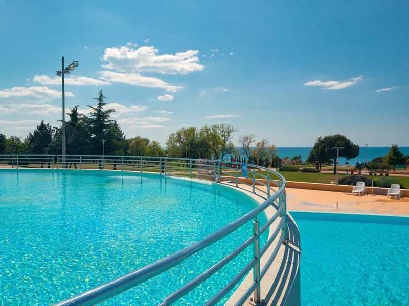 Gran piscina exterior con agua azul clara, rodeada de tumbonas y árboles bajo un cielo azul.