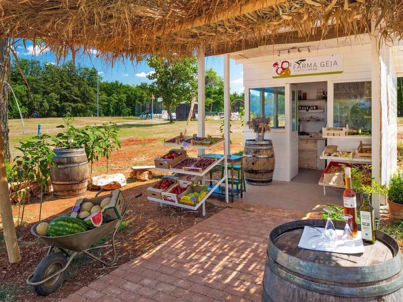 Zona de comida al aire libre con techo de paja, vegetales frescos y barriles decorativos.