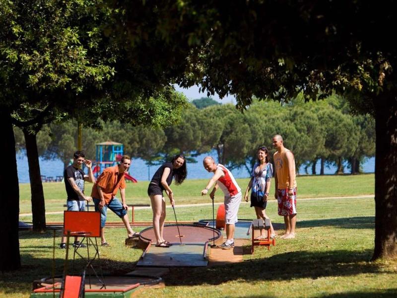 Grupo de personas juega mini golf en un parque cerca de un lago en un día soleado.