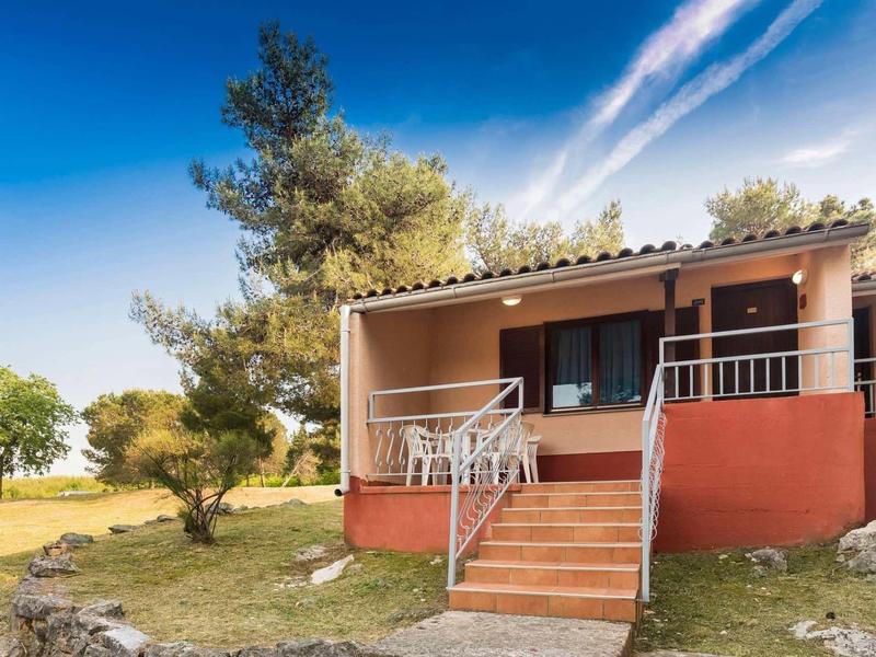 Parterowy domek wakacyjny z tarasem i krzesłami w zielonym otoczeniu pod niebieskim niebem.