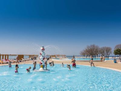 Bambini che giocano in una piscina esterna di un hotel sotto un sole splendente e un cielo azzurro.
